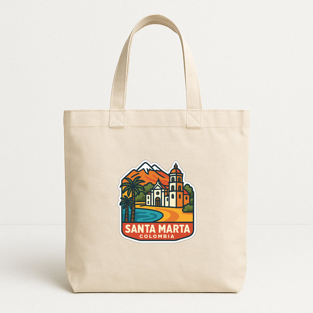 A beige tote bag with a colorful graphic depicting Santa Marta, Colombia, featuring a church, palm trees, and mountains.