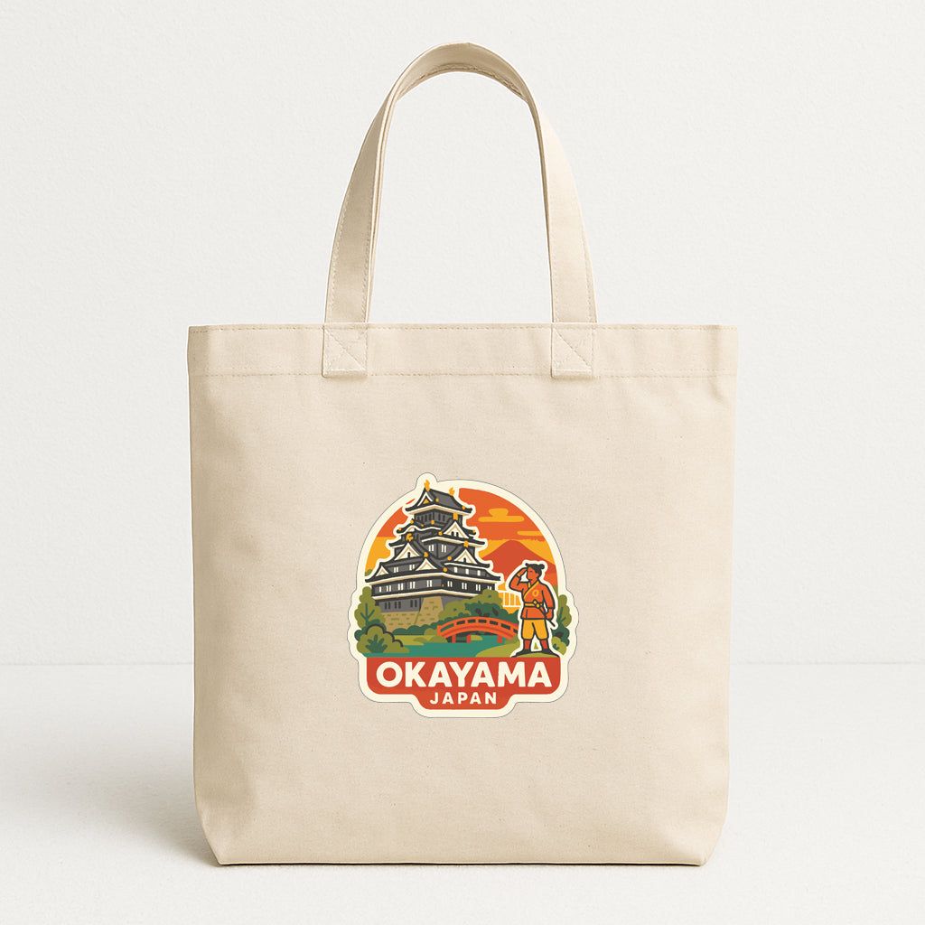 A beige tote bag with a printed design depicting a castle and a person, along with the text "OKAYAMA JAPAN".