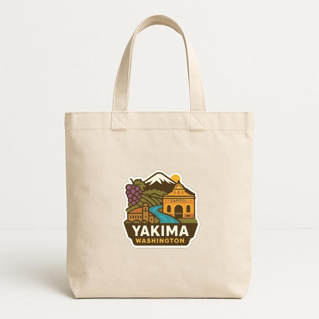 A beige tote bag with a graphic design featuring a mountain, buildings, and a river, along with the text "YAKIMA WASHINGTON".