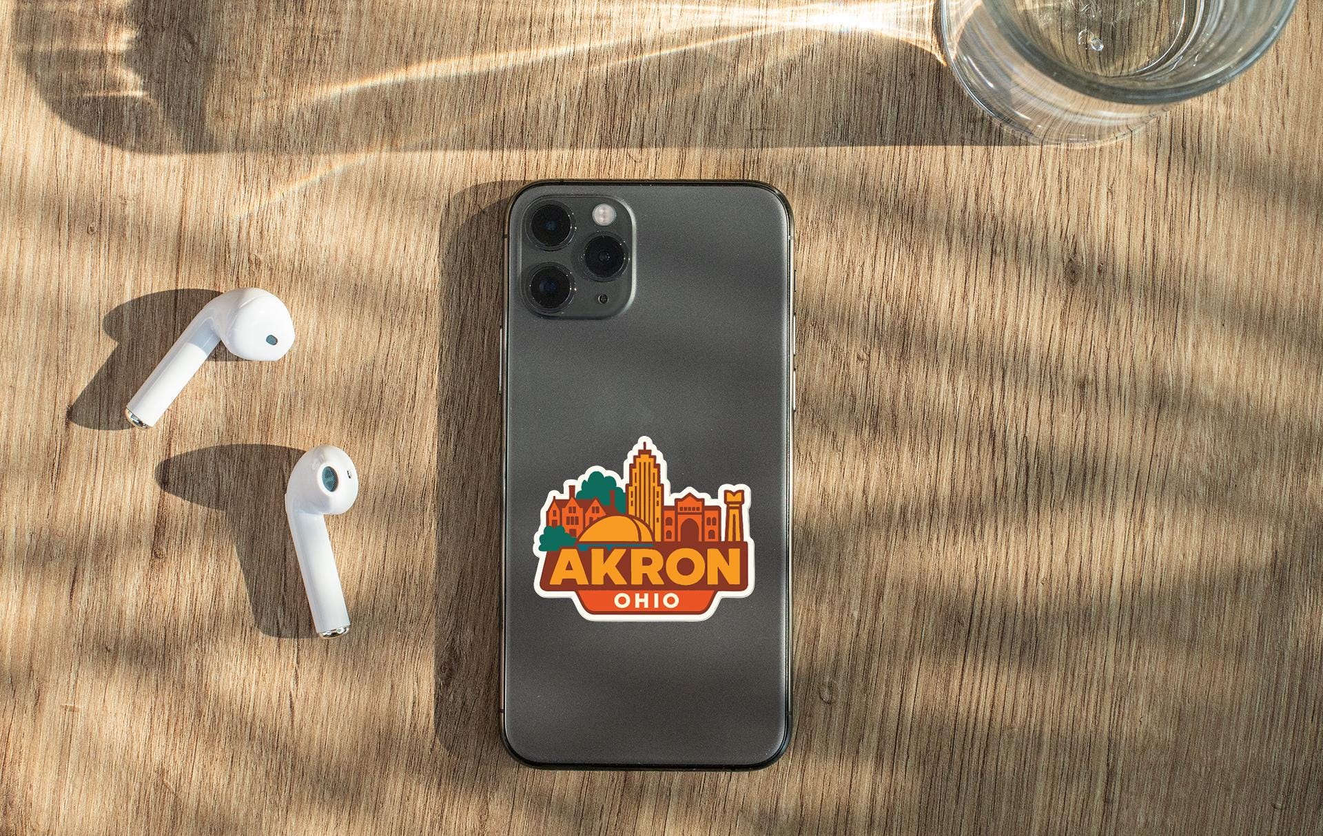 a smartphone with a sticker of the city of Akron, Ohio, placed on a wooden surface alongside a pair of white earbuds and a glass of water.