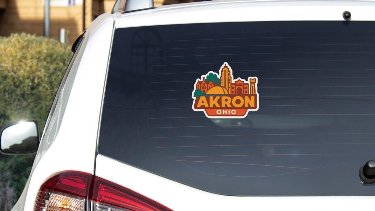 A sticker on the rear window of a white vehicle displaying the city of Akron, Ohio.