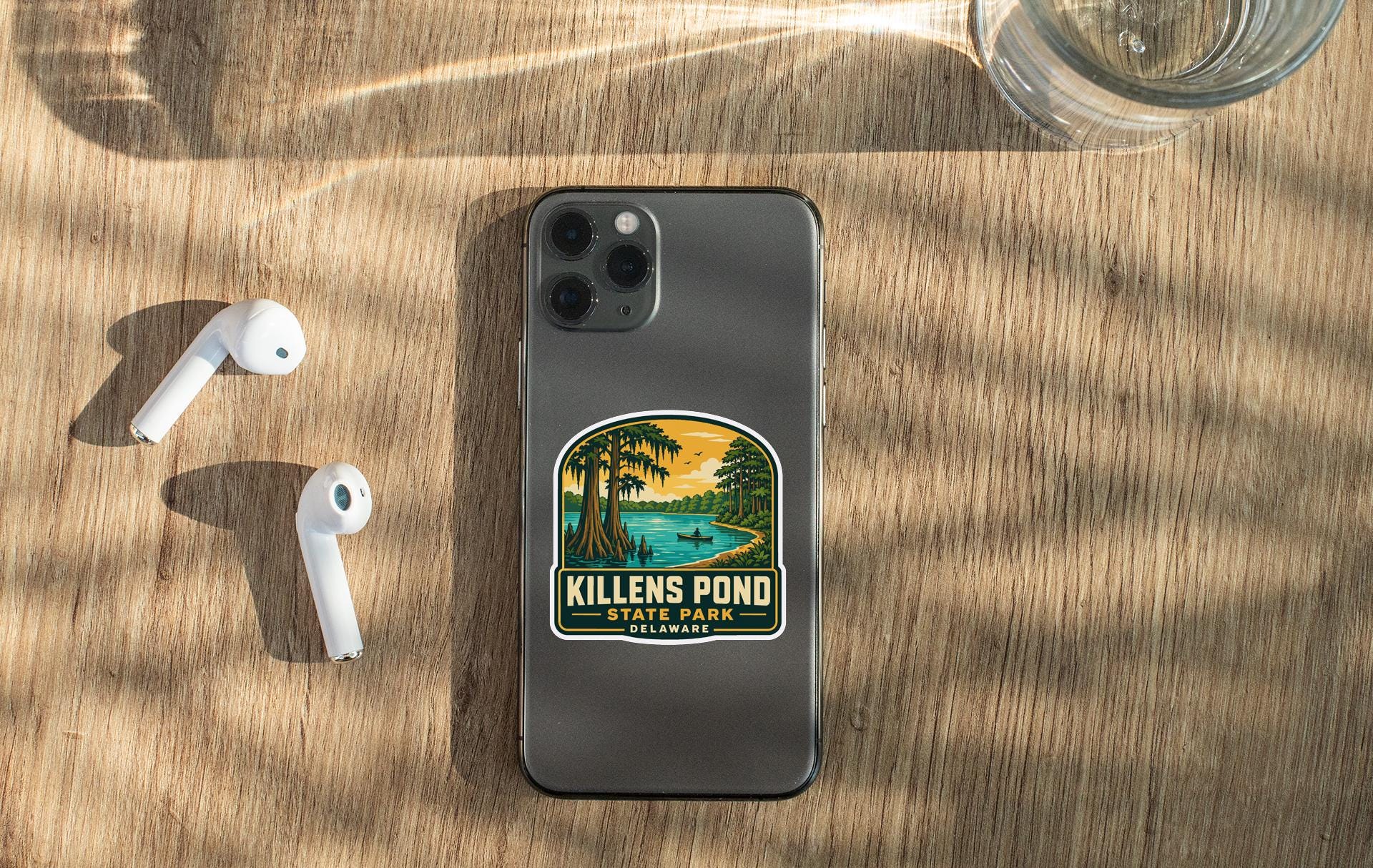 a smartphone with a sticker on the back, placed on a wooden surface alongside a pair of white earbuds and a glass of water.