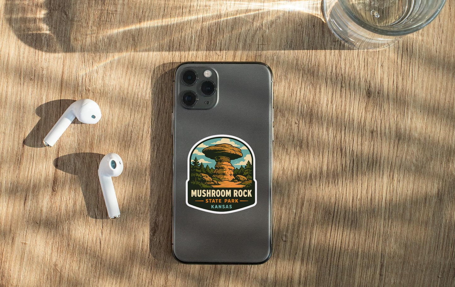 a smartphone with a mushroom rock sticker on the back, placed on a wooden surface alongside a pair of white earbuds and a glass of water.