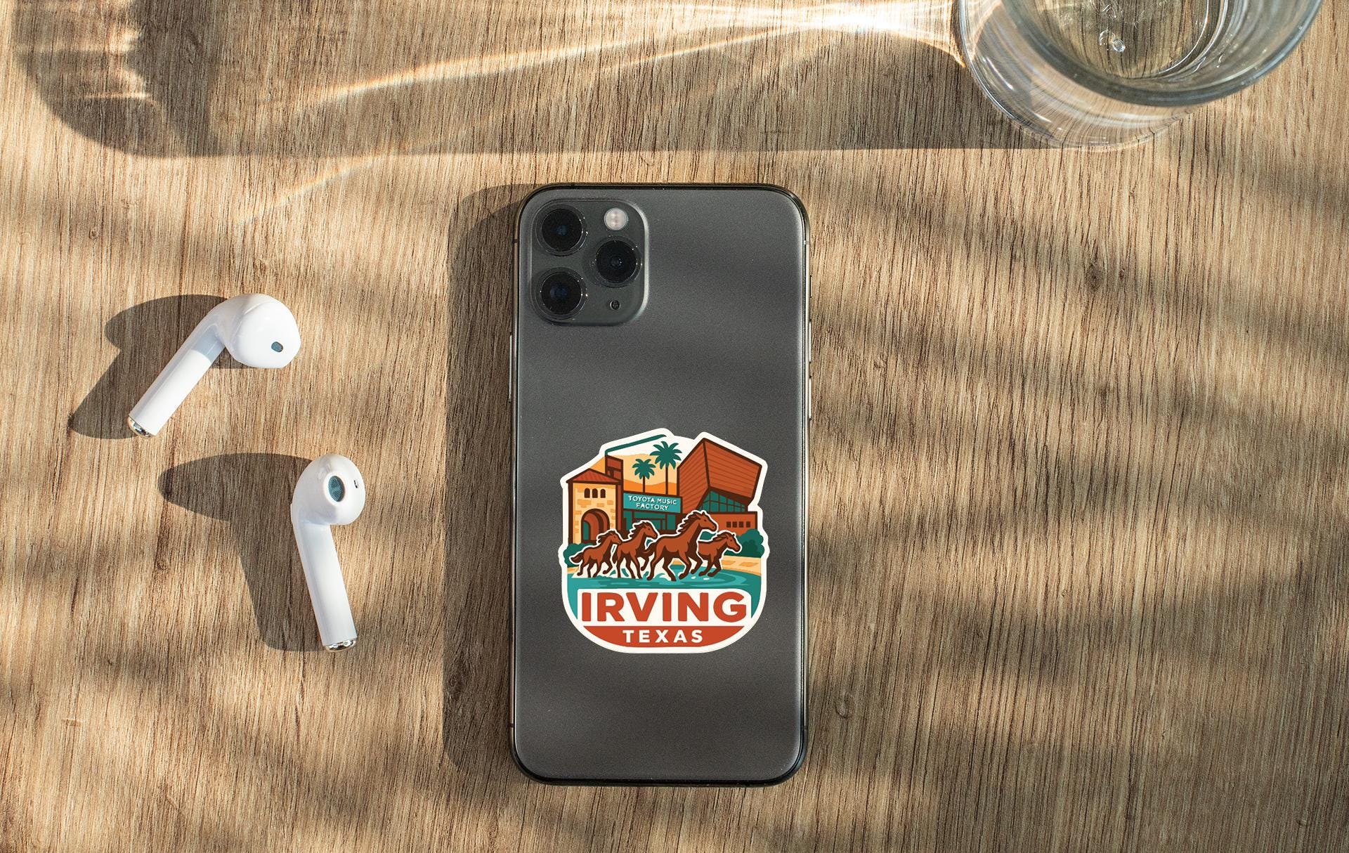 a smartphone with a sticker on the back, placed on a wooden surface alongside a pair of white earbuds and a glass of water.