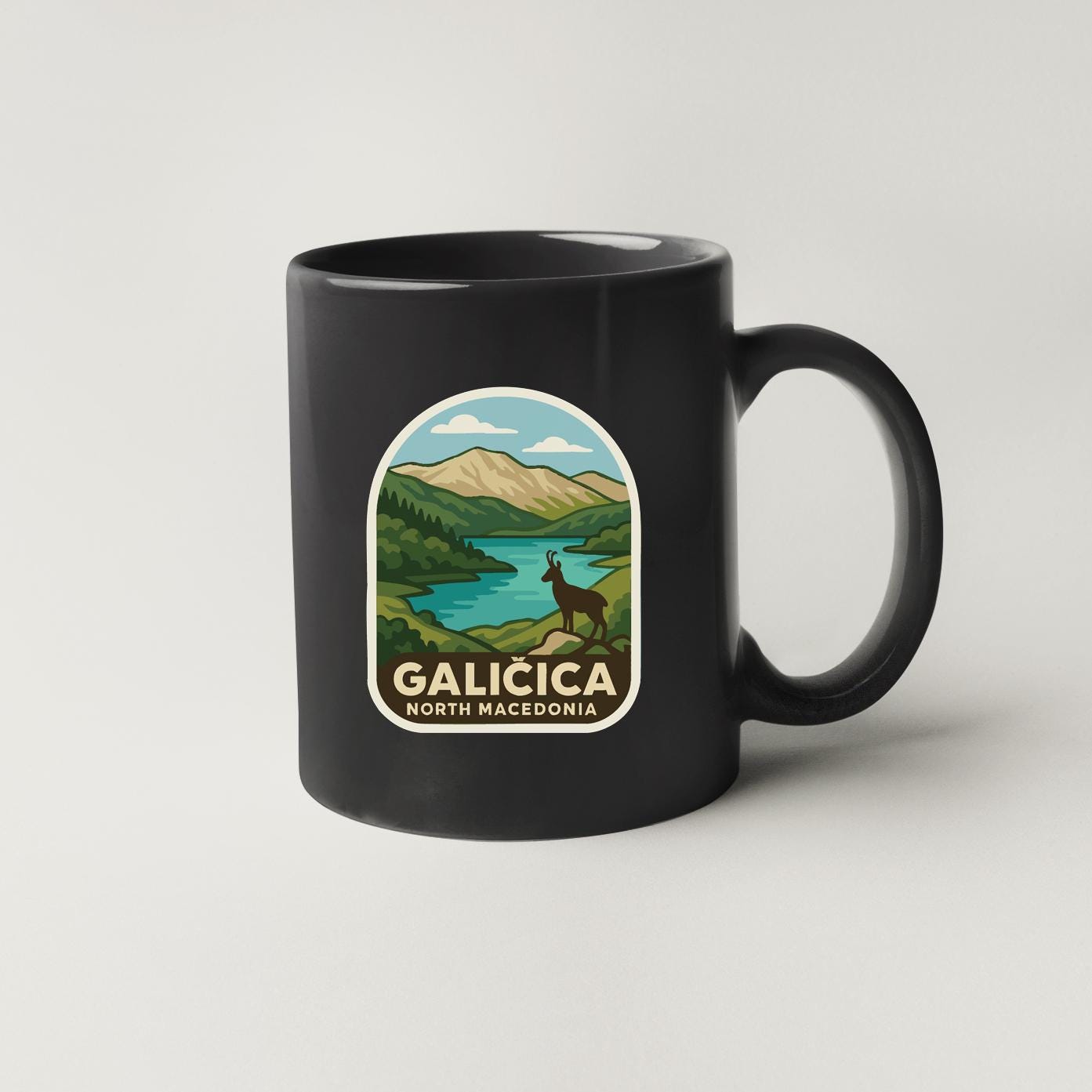 A black coffee mug with a graphic design depicting a scenic landscape with mountains, a lake, and a deer.