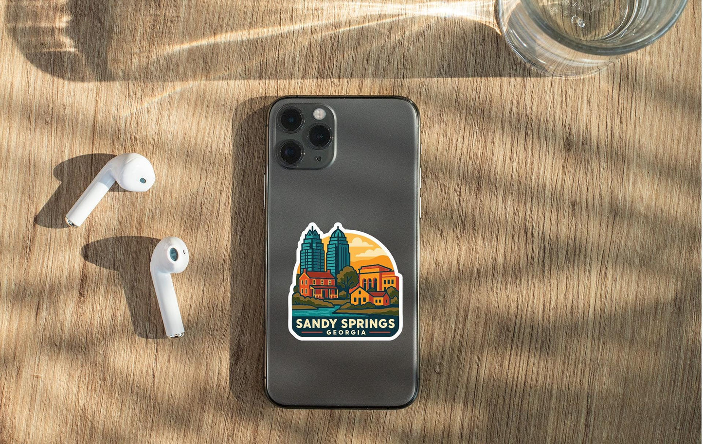 a smartphone with a sticker of a cityscape on it, placed on a wooden surface alongside a pair of white earbuds and a glass of water.