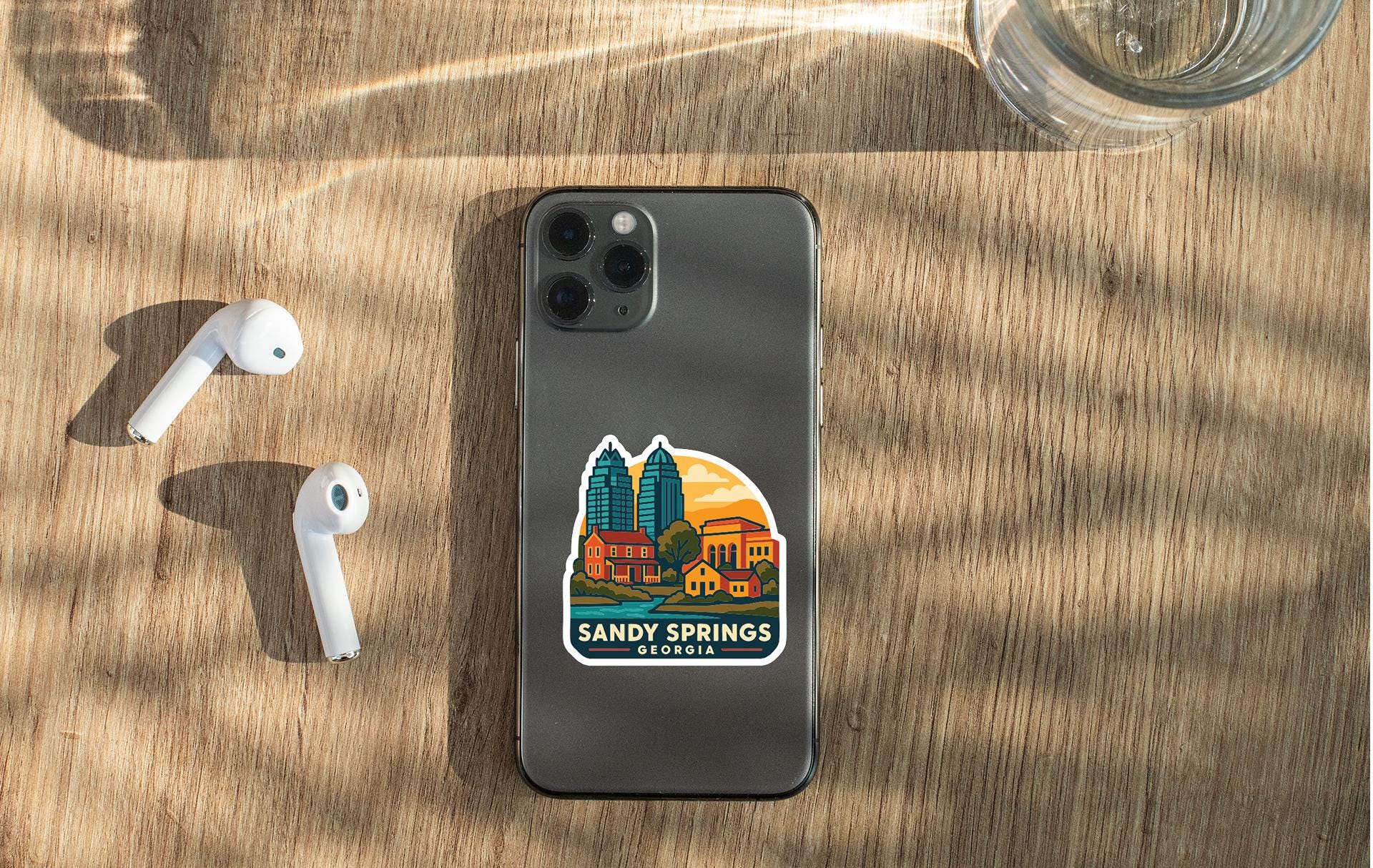 a smartphone with a sticker of a cityscape on it, placed on a wooden surface alongside a pair of white earbuds and a glass of water.