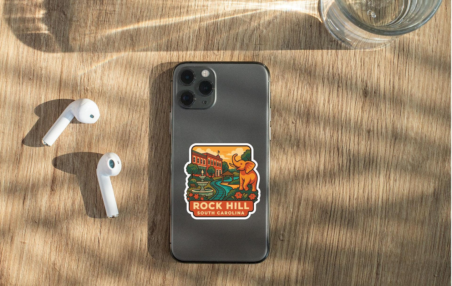 a smartphone with a sticker on the back, placed on a wooden surface alongside a pair of white earbuds and a glass of water.