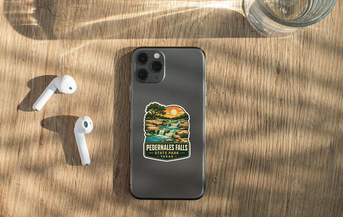 a smartphone with a sticker on the back, placed on a wooden surface alongside a pair of white earbuds and a glass of water.