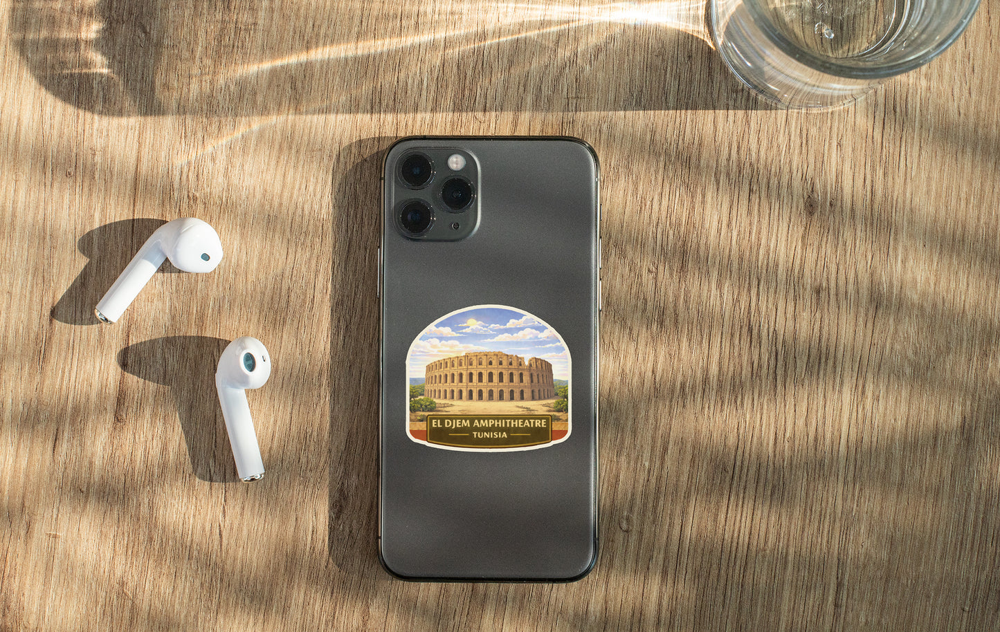 a smartphone with a sticker of the Colosseum on it, placed on a wooden surface alongside a pair of white earbuds and a glass of water.