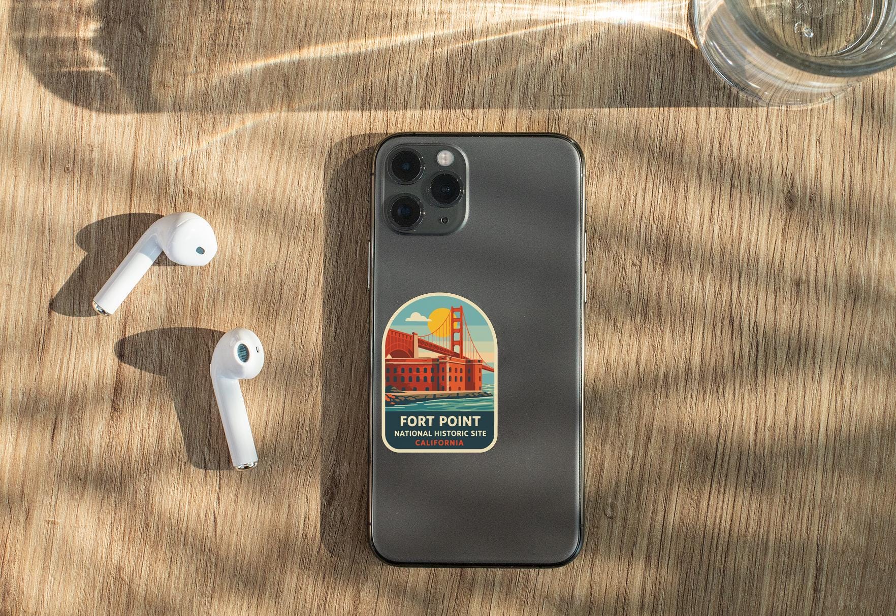 a smartphone with a sticker on the back, placed on a wooden surface alongside a pair of white earbuds and a glass of water.