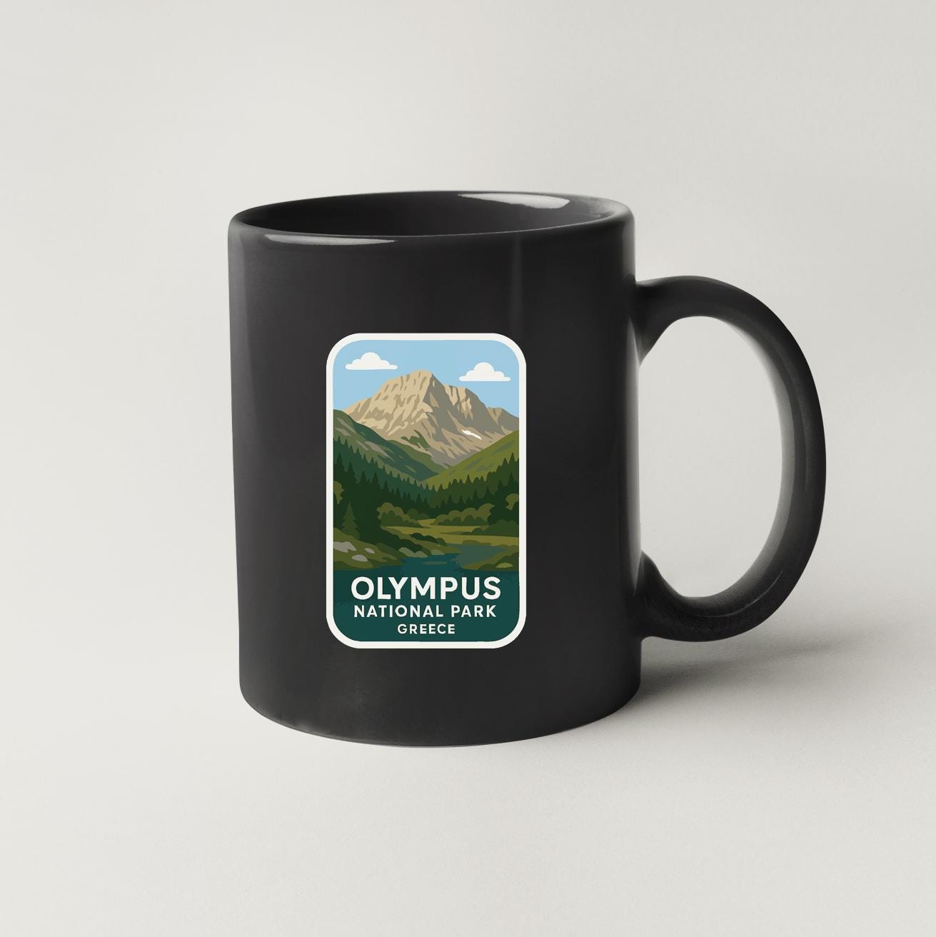 A black coffee mug with a sticker depicting a mountainous landscape and the text "OLYMPUS NATIONAL PARK GREECE".