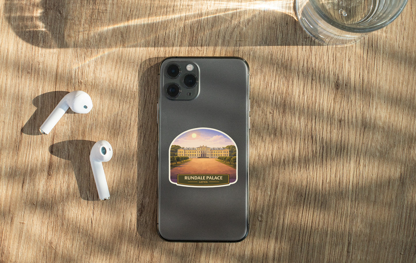 a smartphone with a sticker of the Rundale Palace on its back, along with a pair of white earbuds and a glass of water on a wooden surface.