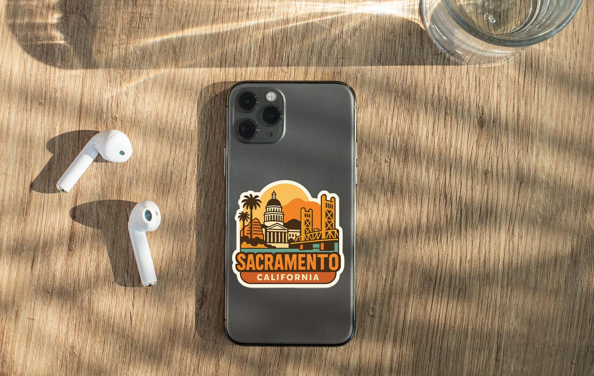 a smartphone with a sticker of the Sacramento, California skyline, along with a pair of white earbuds and a glass of water on a wooden surface.