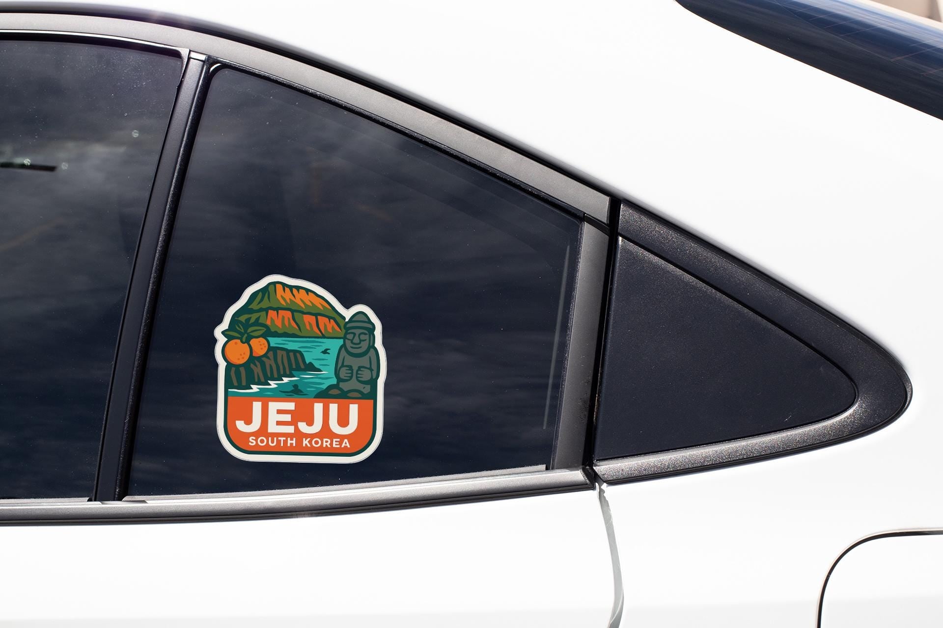 A sticker on the side of a car window, depicting a scenic landscape with mountains and a river, along with the text "JEJU SOUTH KOREA".