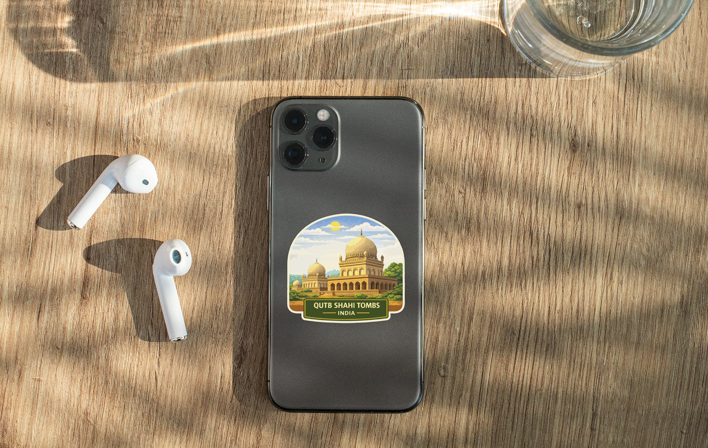 a smartphone with a sticker of a mosque on its back, along with a pair of white earbuds and a glass of water on a wooden surface.