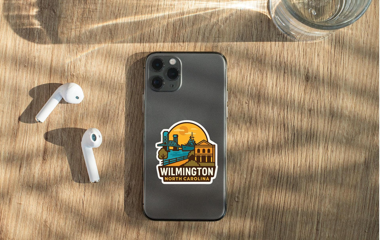 a smartphone with a sticker of Wilmington, North Carolina, placed on a wooden surface alongside a pair of white earbuds and a glass of water.