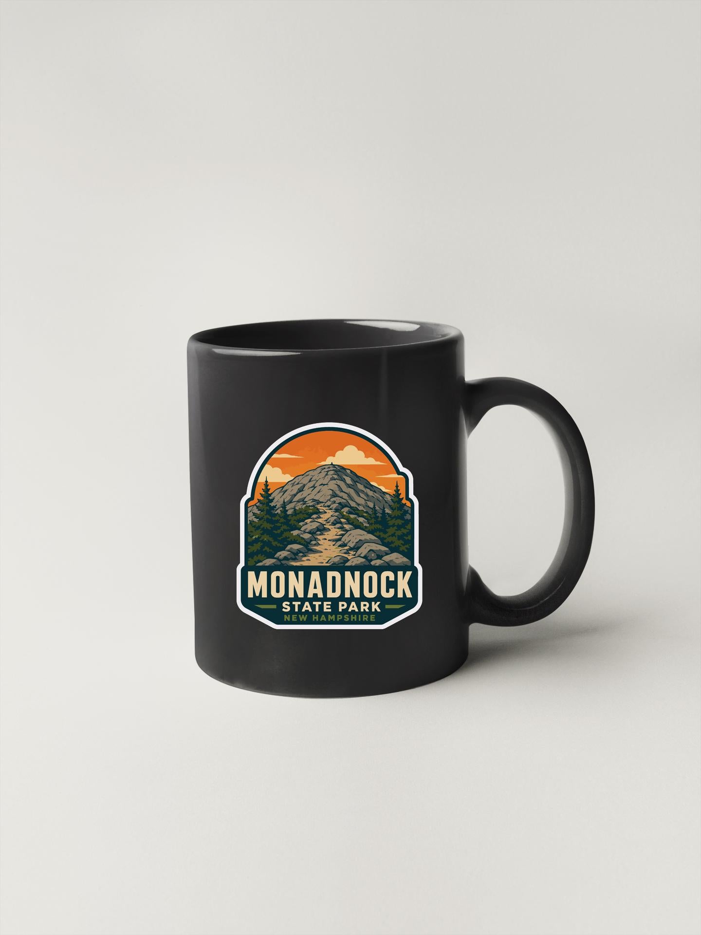 A black coffee mug with a graphic design featuring a mountain landscape and the text "MONADNOCK STATE PARK NEW HAMPTON, N.C."