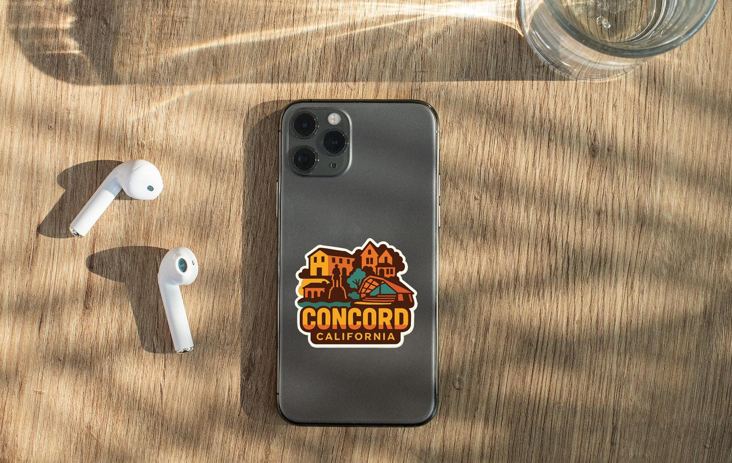 a smartphone with a sticker on it, placed on a wooden surface alongside a pair of white earbuds and a glass of water.