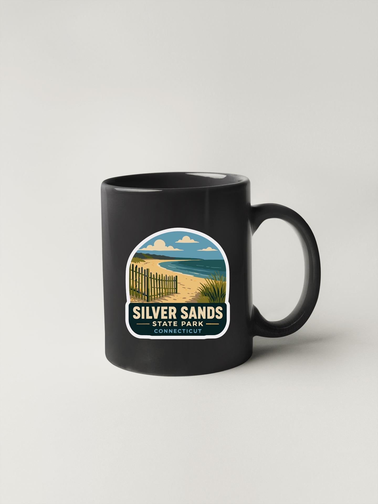 A black coffee mug with a graphic design depicting a beach scene and a fence, along with the text "SILVER SAND'S STATE PARK CONNECTICUT".