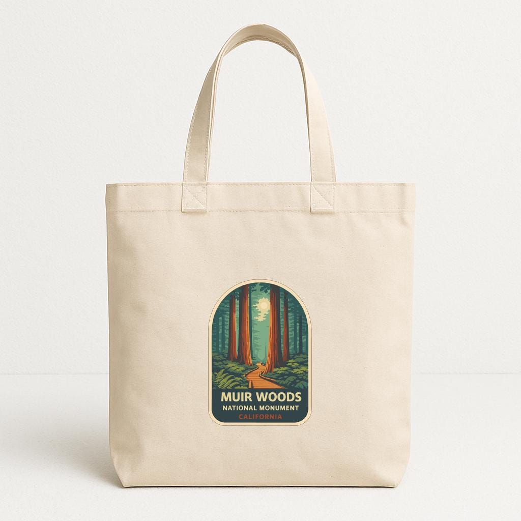 A beige tote bag with a printed label that says "MUR WOODS NATIONAL MONUMENT CALIFORNIA" and features an illustration of a forest with tall trees and a path leading through the trees.
