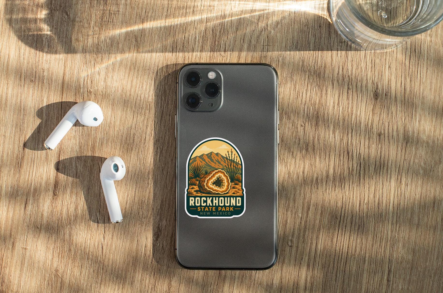 a smartphone with a sticker on it, placed on a wooden surface alongside a pair of white earbuds and a glass of water.