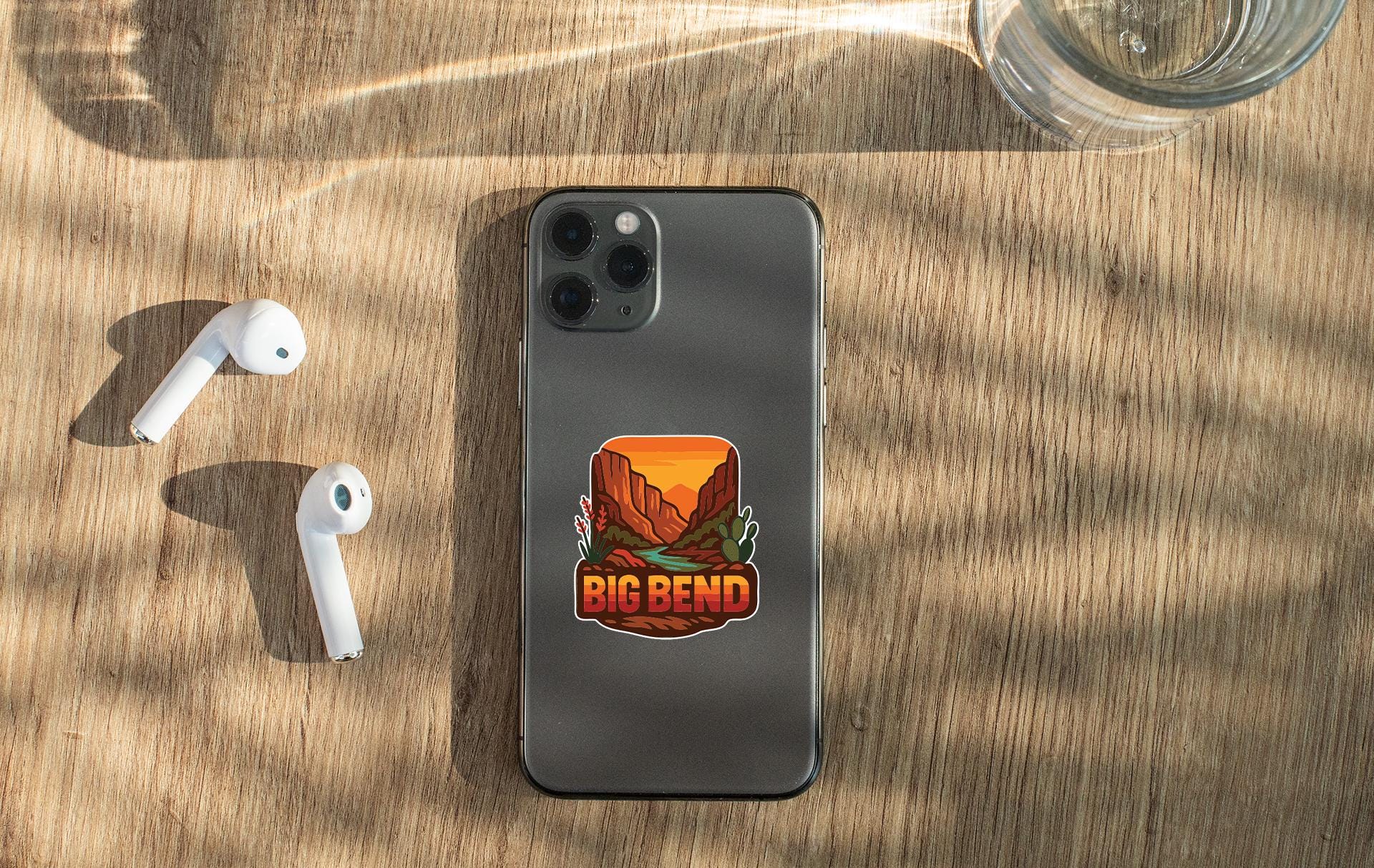 a smartphone with a sticker on the back, along with a pair of white earbuds and a glass of water on a wooden surface.
