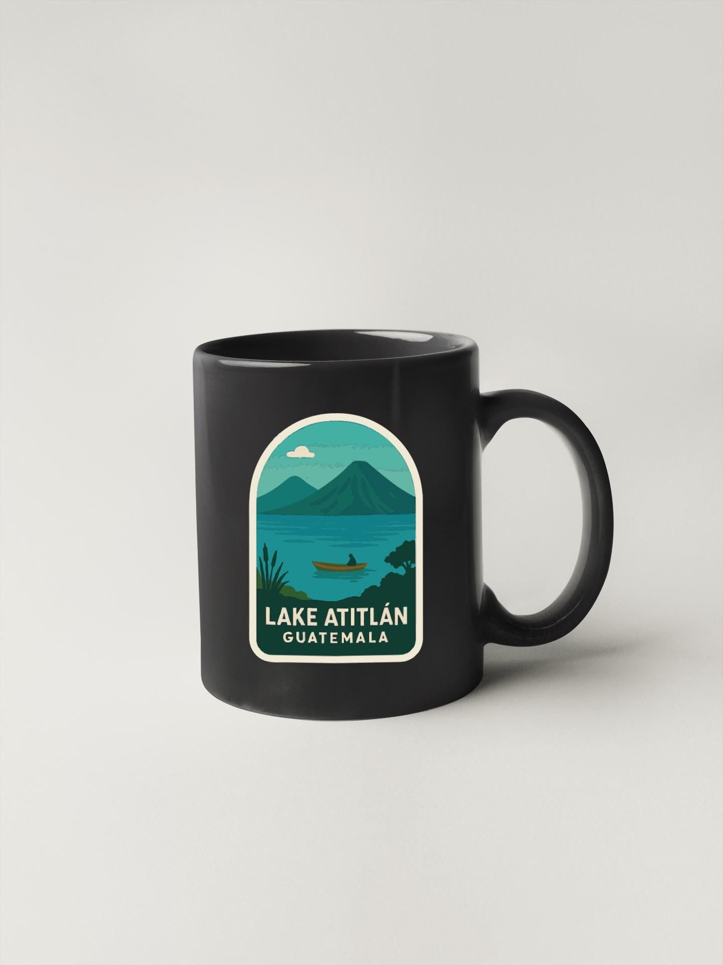 A black coffee mug with a graphic design depicting a lake, mountains, and a boat, along with the text "LAKÉ ATITLAN QUAYTEMAL".