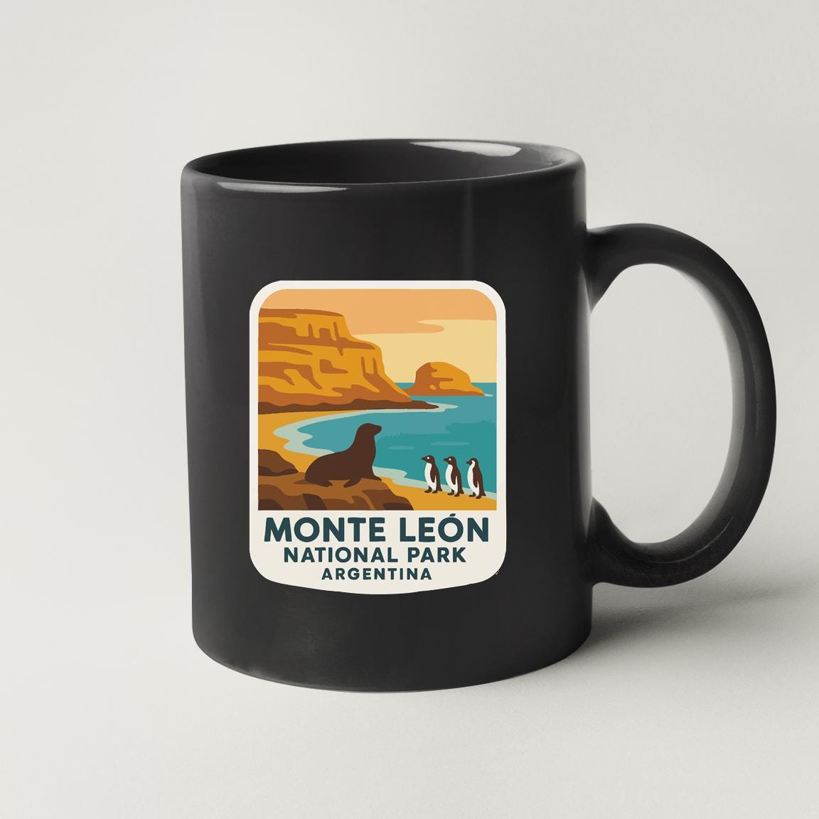 A black coffee mug with a label that features an illustration of a seal on a rocky shoreline with penguins, along with the text "MONTE LEON NATIONAL PARK ARGENTINA".