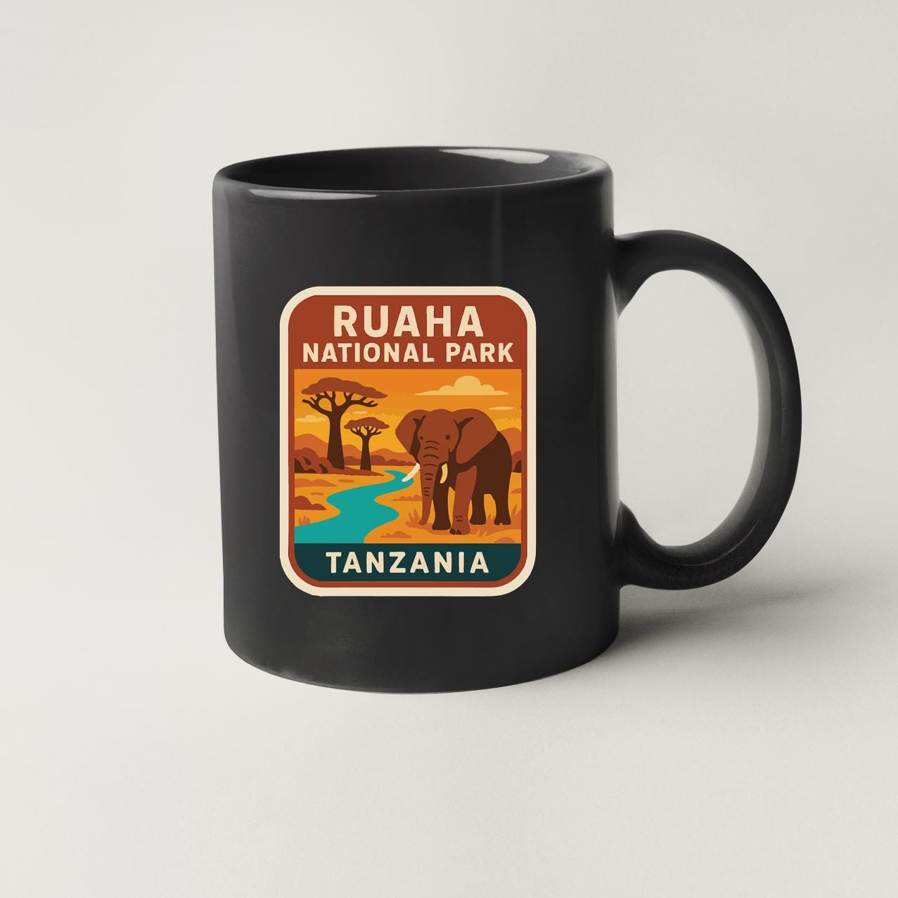 A black coffee mug with a colorful illustration of an elephant and a river, along with the text "RUAHAN NATIONAL PARK TANZANIA".