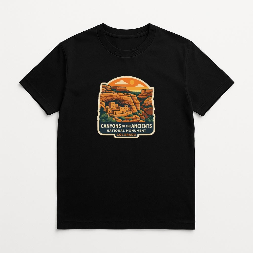 A black t-shirt with a graphic design depicting a desert landscape with a cliff and a cave, along with the text "CANYONS OF THE ANCIENTS NATIONAL PARK COLORADO".