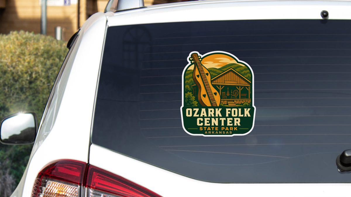 A sticker on the rear window of a white vehicle, featuring an illustration of a cabin and the text "Ozark Folk Center State Park Arkansas".