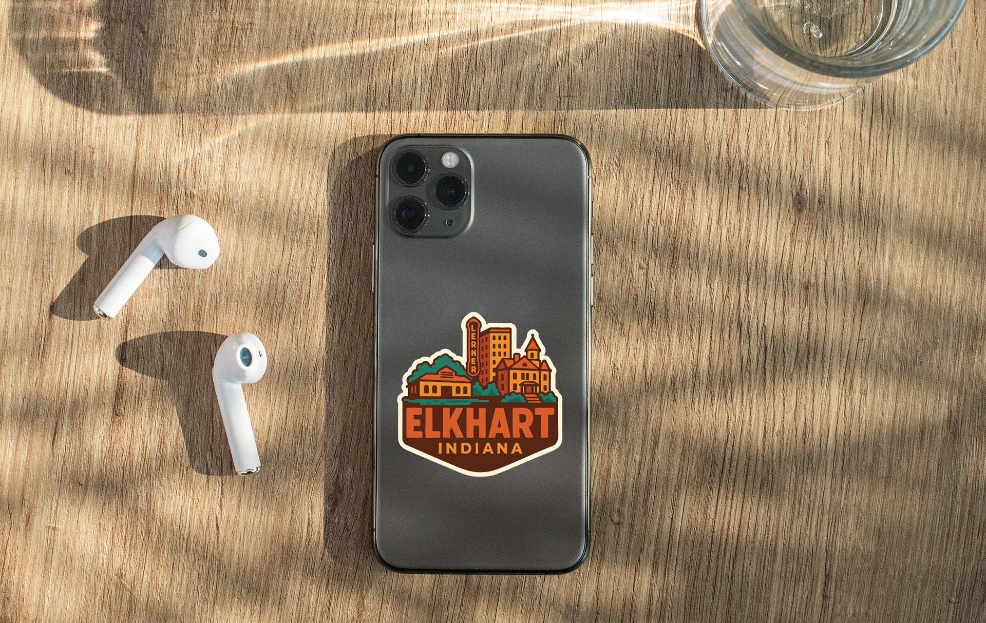 a smartphone with a sticker on the back, placed on a wooden surface alongside a pair of white earbuds and a glass of water.