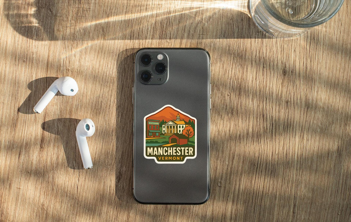 a smartphone with a sticker on the back, placed on a wooden surface alongside a pair of white earbuds and a glass of water.