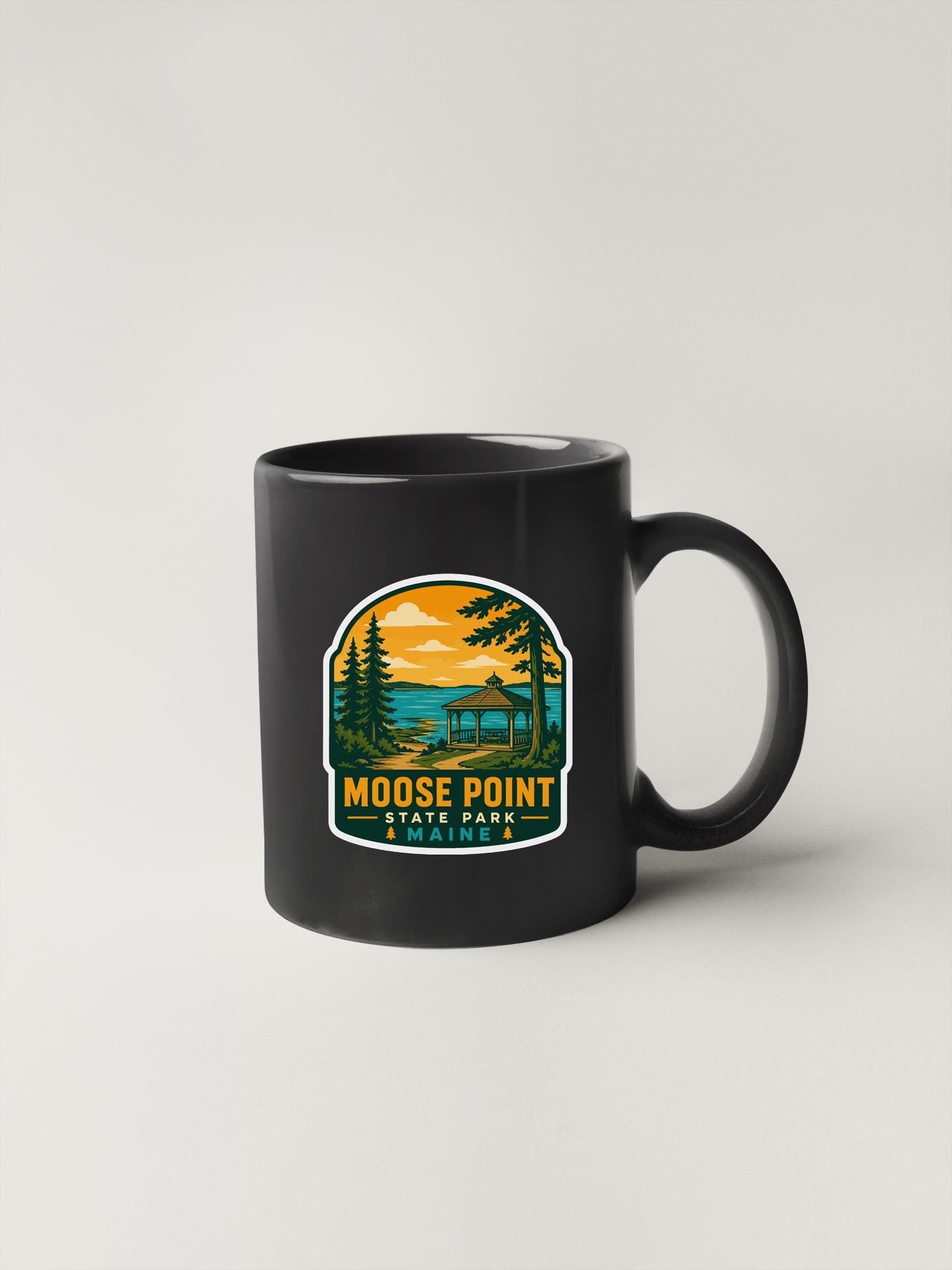 A black coffee mug with a graphic design depicting a scenic landscape with a lake, trees, and a sunset.