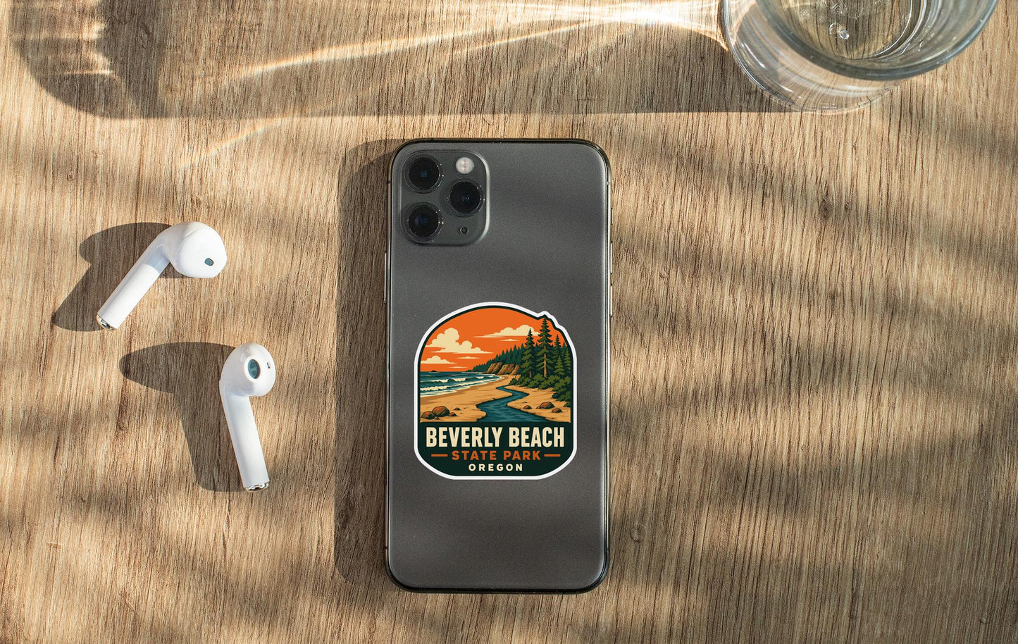 a smartphone with a sticker of Beverly Beach, Oregon, placed on a wooden surface alongside a pair of white earbuds and a glass of water.