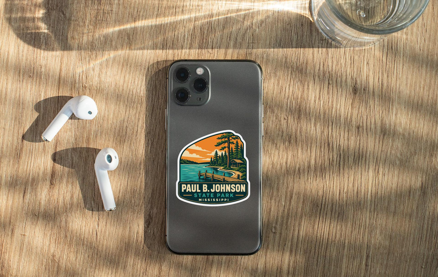 a smartphone with a sticker on the back, placed on a wooden surface alongside a pair of white earbuds and a glass of water.
