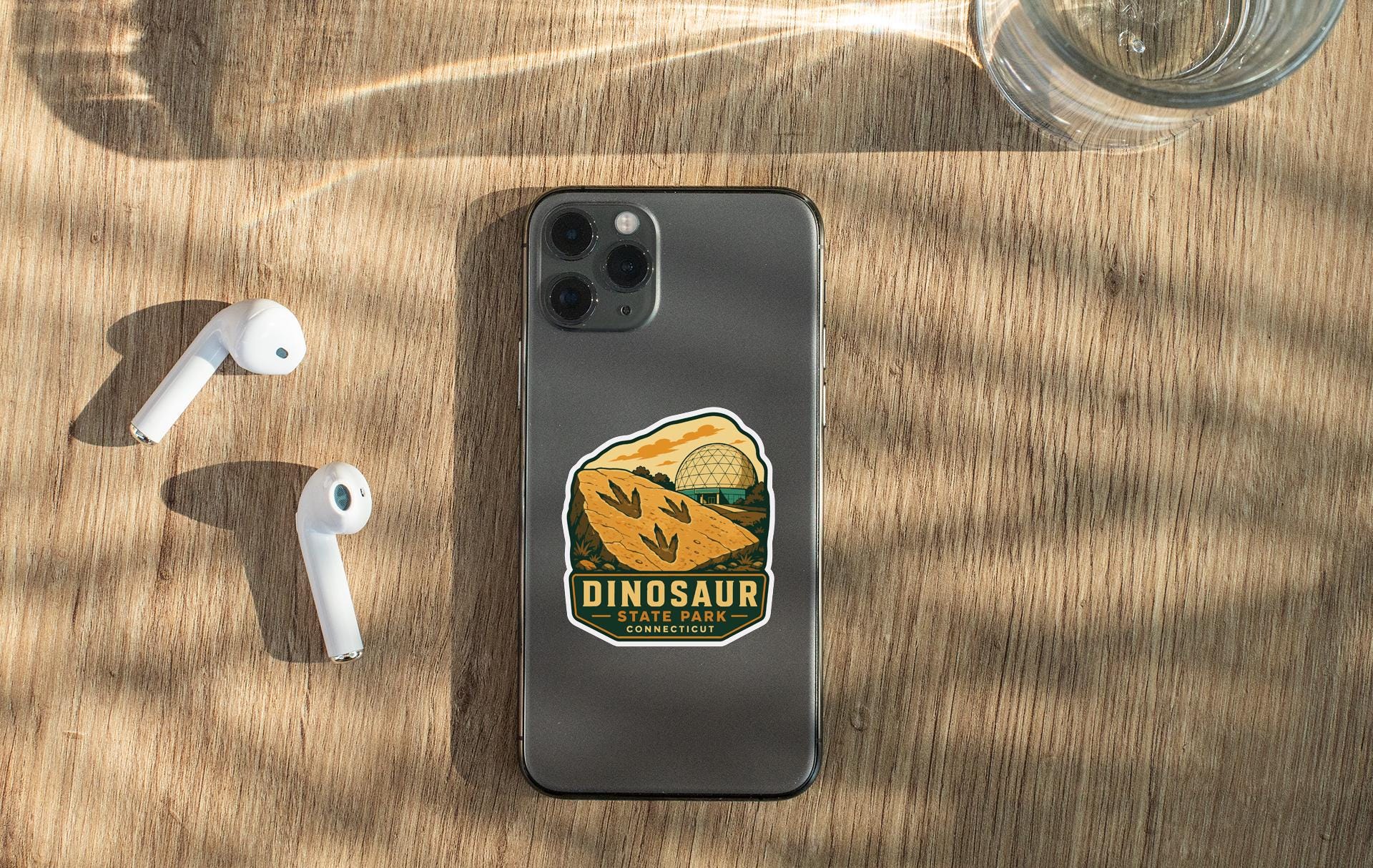a smartphone with a sticker of Dinosaur State Park on it, along with a pair of white earbuds and a glass of water on a wooden surface.
