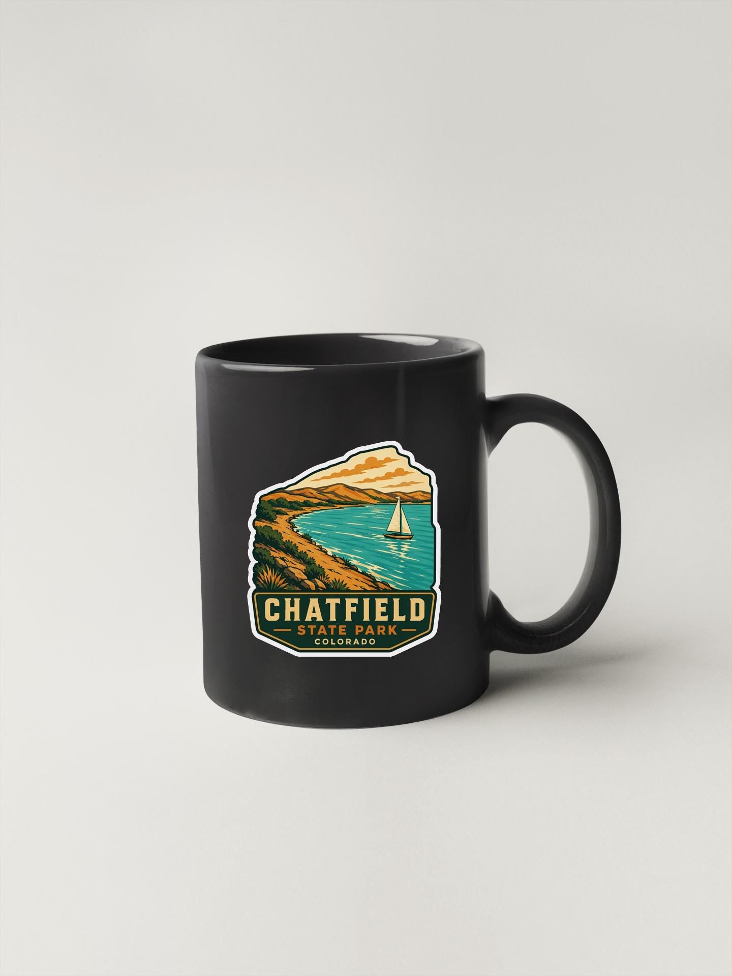 A black coffee mug with a graphic design depicting a scenic landscape with a sailboat on a lake.