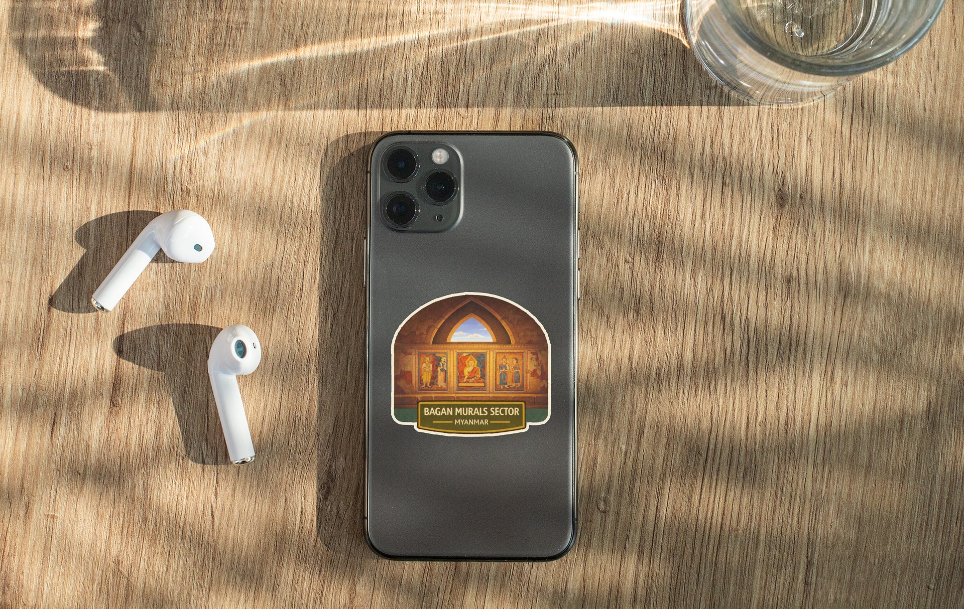 a smartphone with a sticker on the back, placed on a wooden surface alongside a pair of white earbuds and a glass of water.