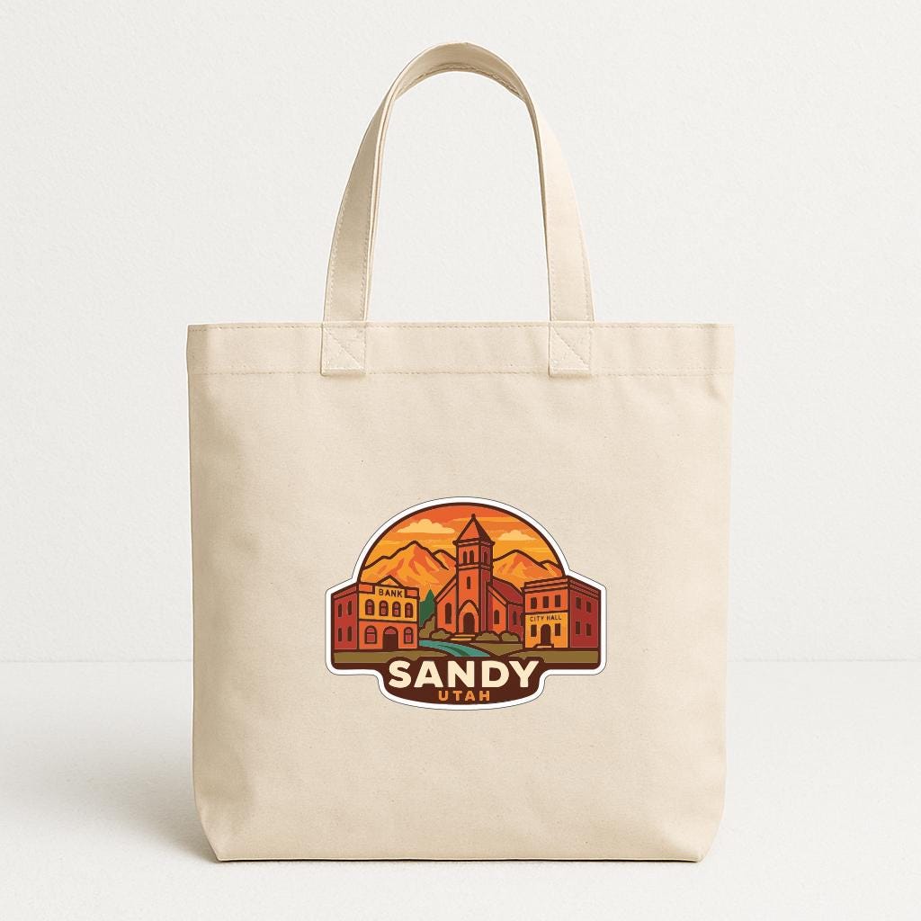 A beige tote bag with a graphic design depicting a cityscape and the text "SANDY UTAH" printed on it.