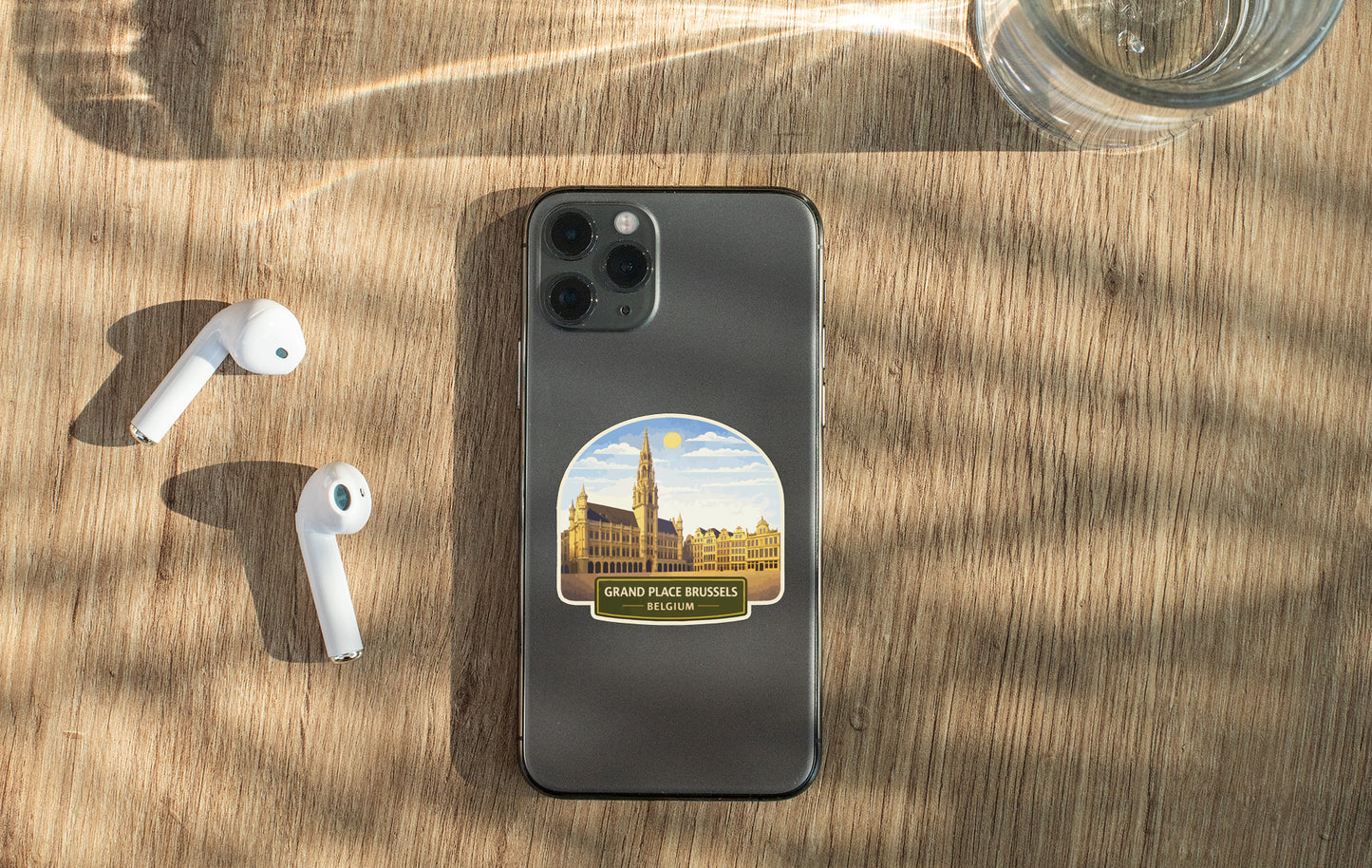 a smartphone with a sticker of Brussels, Belgium, placed on a wooden surface alongside a pair of white earbuds and a glass of water.