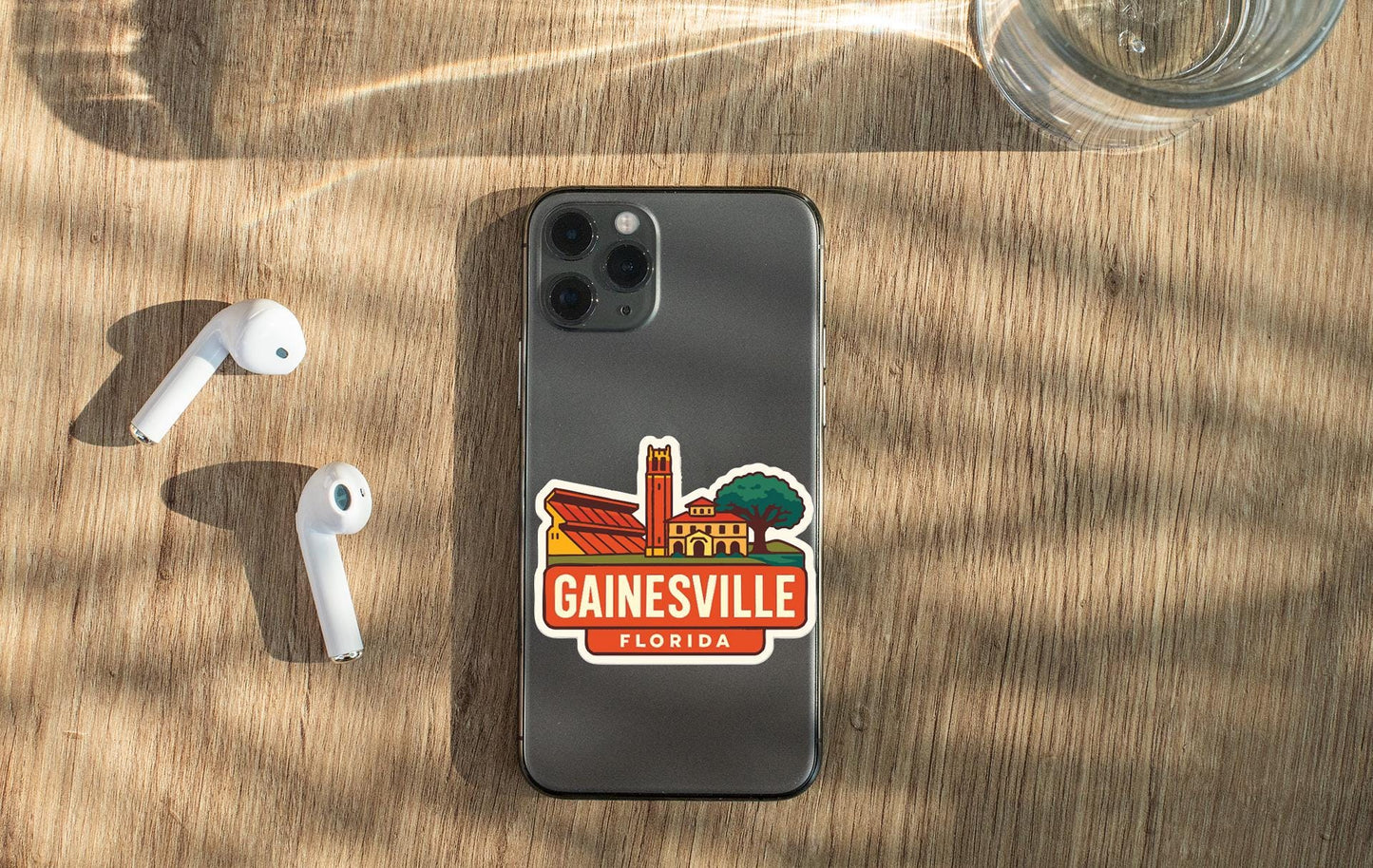 a smartphone with a sticker of Gainesville, Florida, placed on a wooden surface alongside a pair of white earbuds and a glass of water.