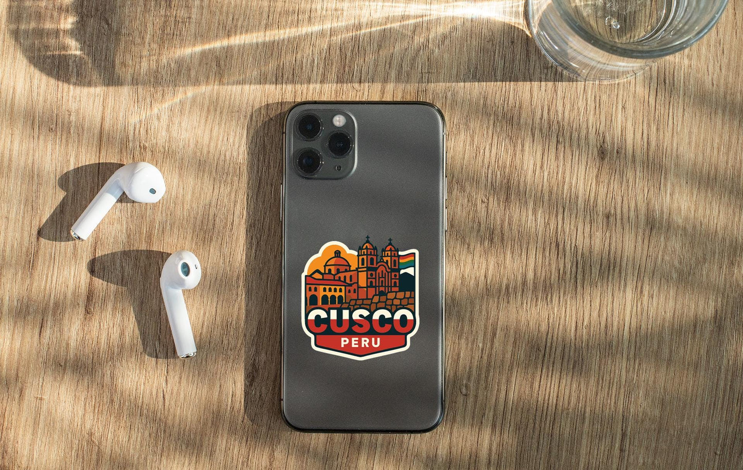 a smartphone with a sticker of a city on it, placed on a wooden surface alongside a pair of white earbuds and a glass of water.