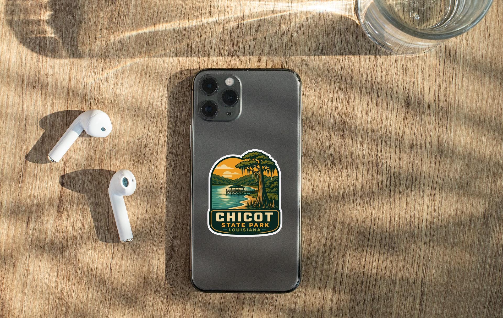 a smartphone with a sticker on the back, placed on a wooden surface alongside a pair of white earbuds and a glass of water.