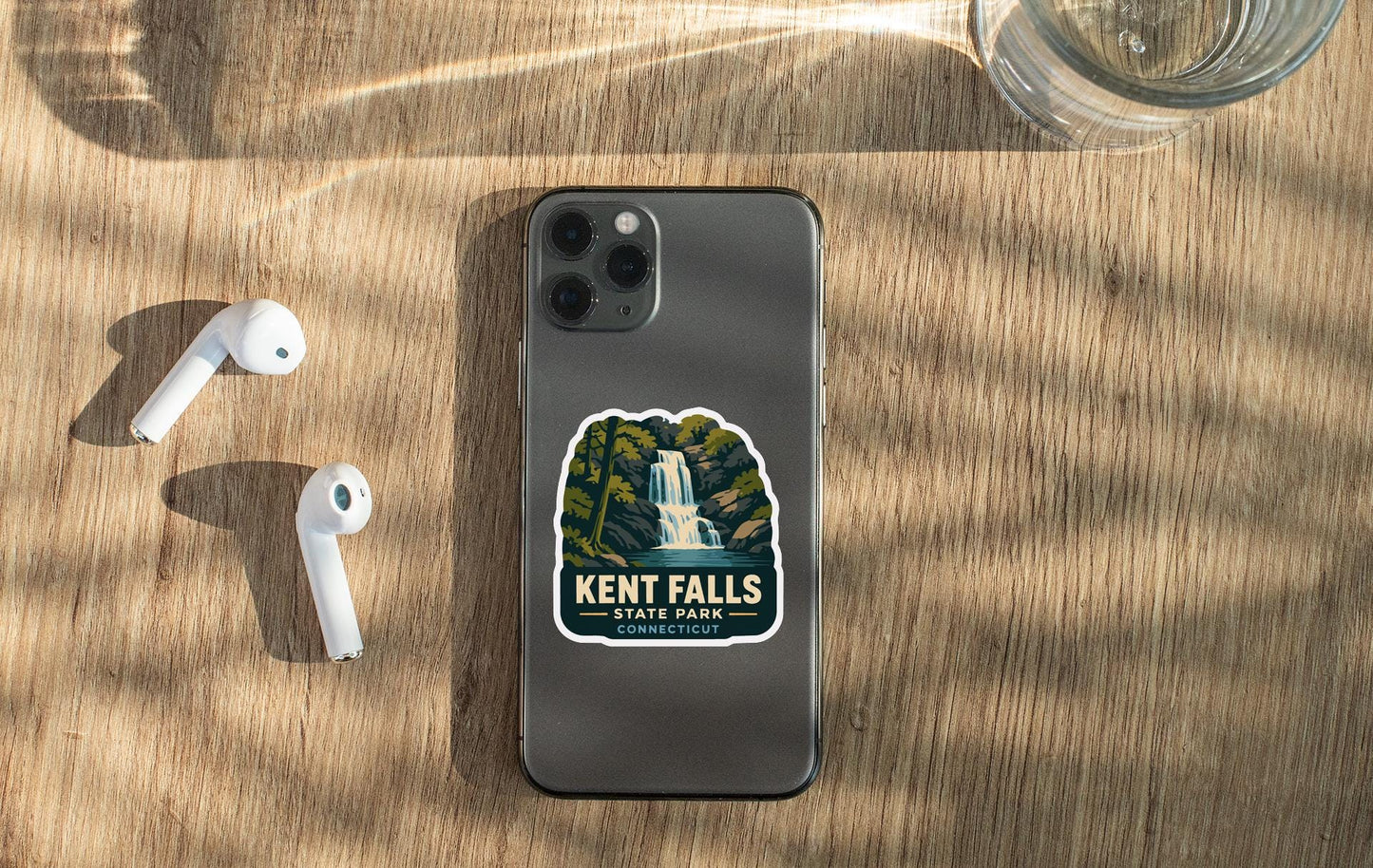 a smartphone with a Kent Falls State Park connection sticker on it, placed on a wooden surface alongside a pair of white earbuds and a glass of water.
