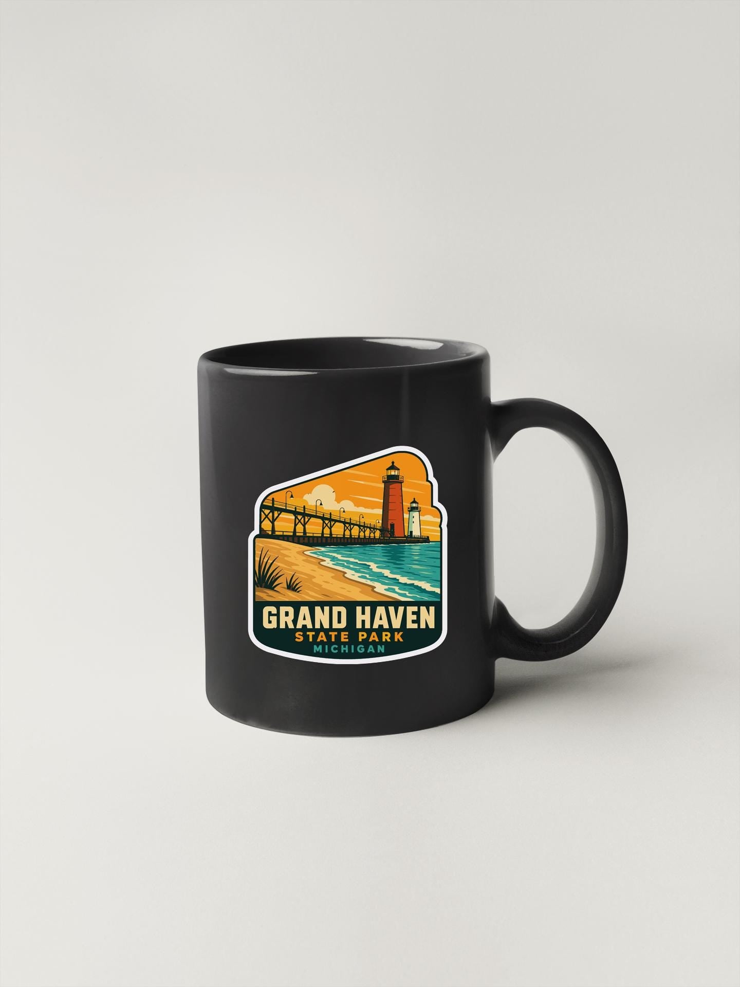 A black coffee mug with a colorful graphic depicting a lighthouse and a beach scene, along with the text "GRAND HAVEN STATE PARK".