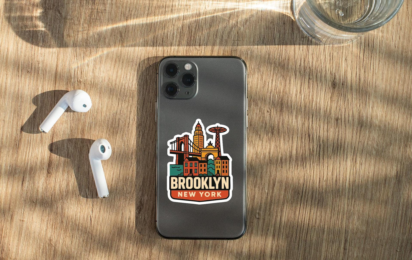 a smartphone with a sticker of the Brooklyn skyline on it, placed on a wooden surface alongside a pair of white earbuds and a glass of water.
