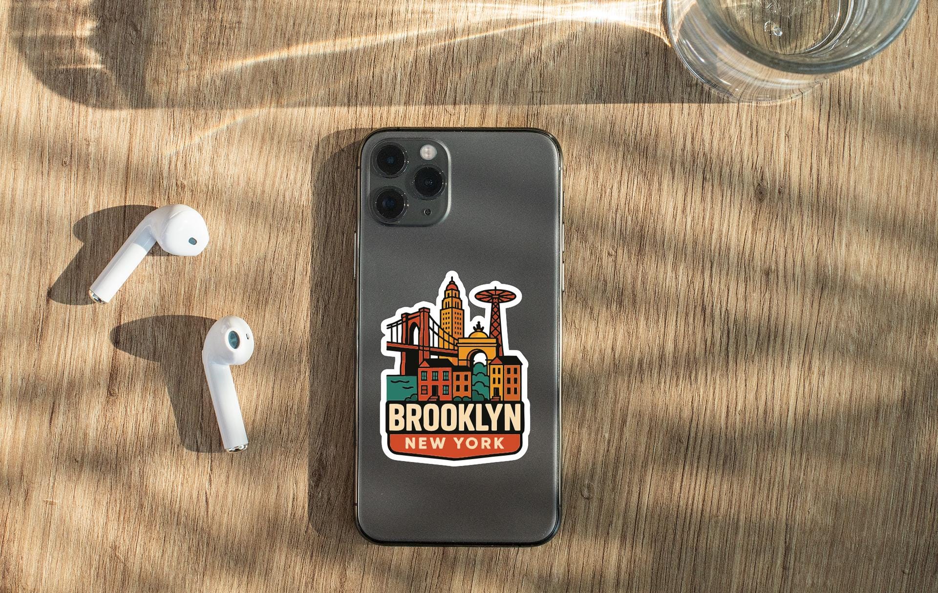 a smartphone with a sticker of the Brooklyn skyline on it, placed on a wooden surface alongside a pair of white earbuds and a glass of water.