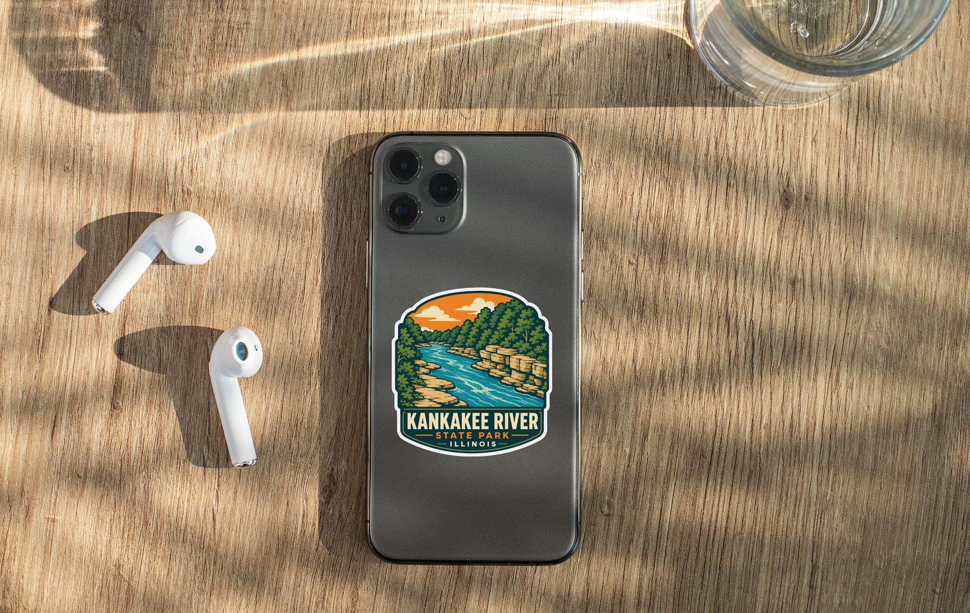 a smartphone with a sticker of the Kankakee River on it, along with a pair of white earbuds and a glass of water on a wooden surface.
